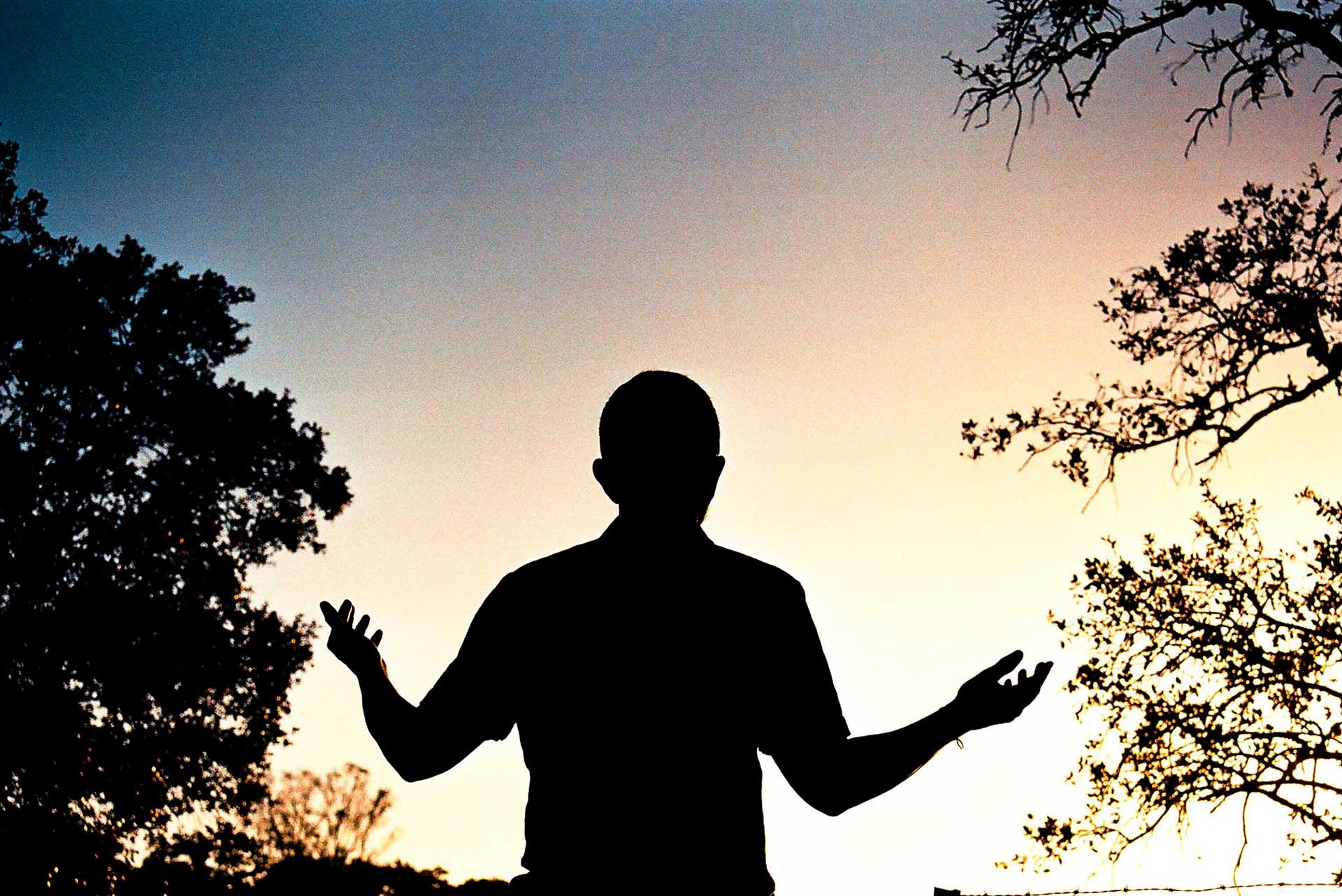 Silueta de hombre de espaldas alza las manos en oración con un fondo de amanecer
