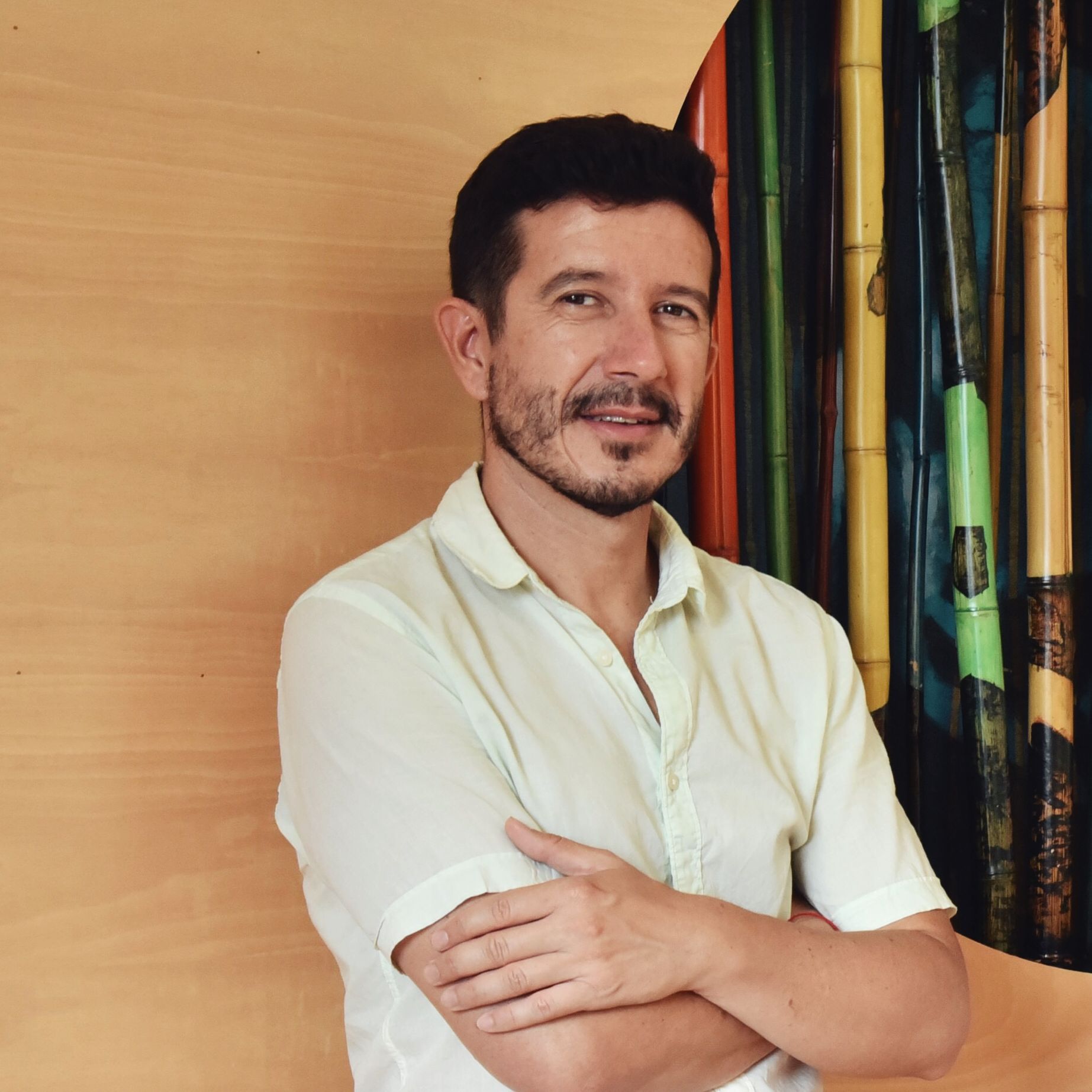 Juan Manuel Palma vestido de color claro mira a la cámara y cruza los brazos sobre un fondo de madera y bambú